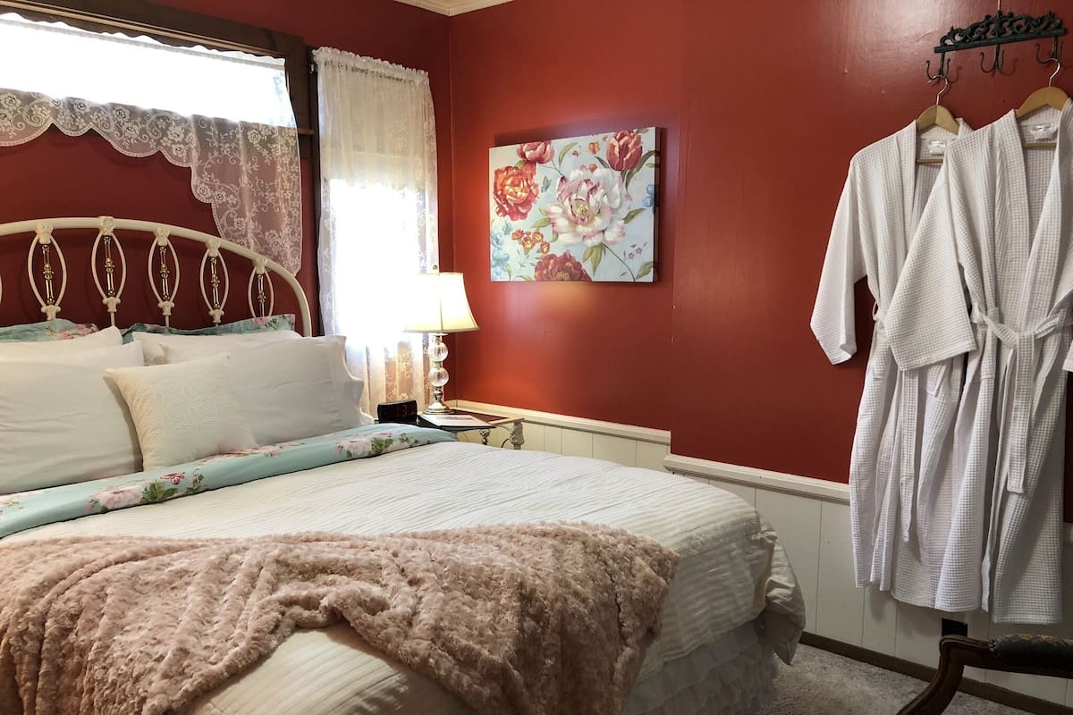 A bedroom with a bed, a nightstand, a lamp, a painting, and a robe hanging on a rack.