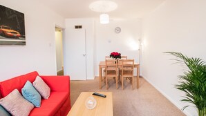 Un salon avec un canapé rouge, une table à manger en bois avec quatre chaises, une petite table avec une télécommande, une plante en pot et un tableau accroché au mur.