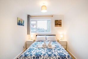 A bedroom with a bed, two bedside lamps, a window with a view, and two wall-mounted artworks. - Apton Court (Bishop's Stortford)