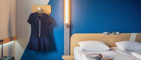 Una habitación de hotel con cama, mesita de noche, luz de pared y perchero con un vestido.