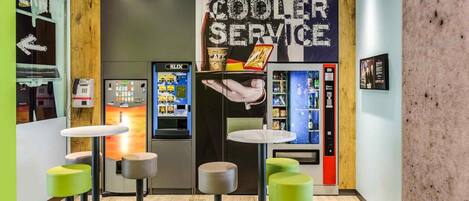 Un área de bar moderna con taburetes, un letrero que dice 'COOLER SERVICE' y máquinas expendedoras.