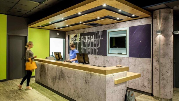 A reception desk with a wooden counter and a marble wall. - Ibis Budget Salzburg Airport (Wals-Siezenheim)