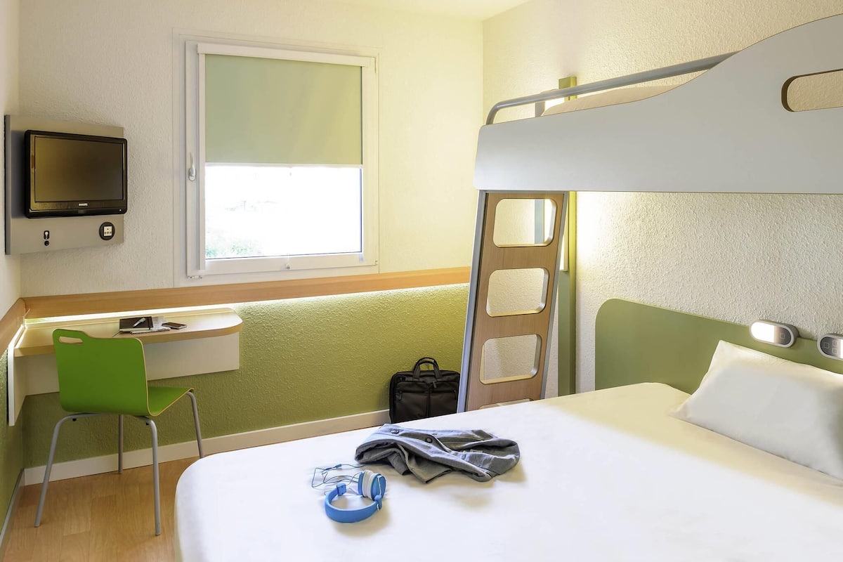 Une chambre d’hôtel avec un lit superposé, un bureau avec une chaise, une télévision et une fenêtre.