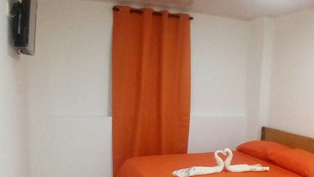 Un lit simple avec une couvre-lit orange et des coussins assortis, un téléviseur blanc fixé au mur et une fenêtre avec des rideaux orange.