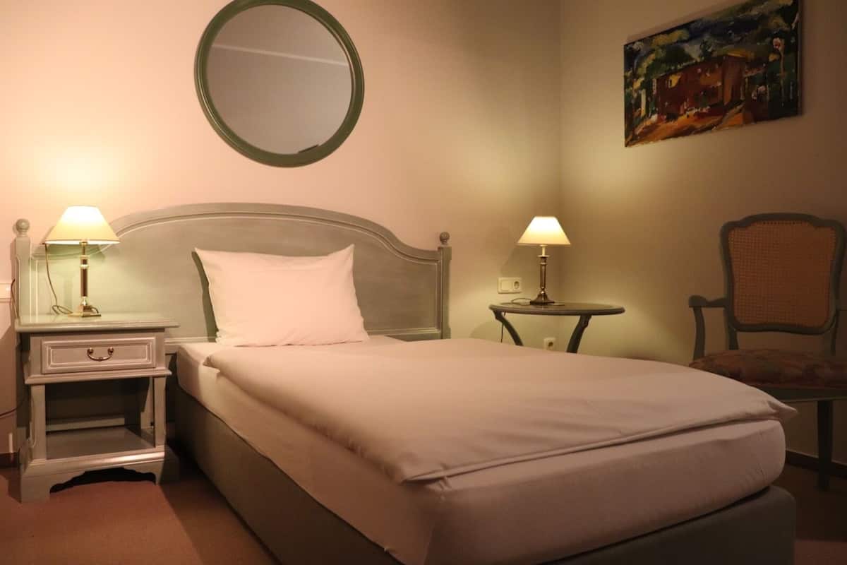 A hotel room with a bed, two bedside tables with lamps, a round mirror, a chair, and a painting on the wall.