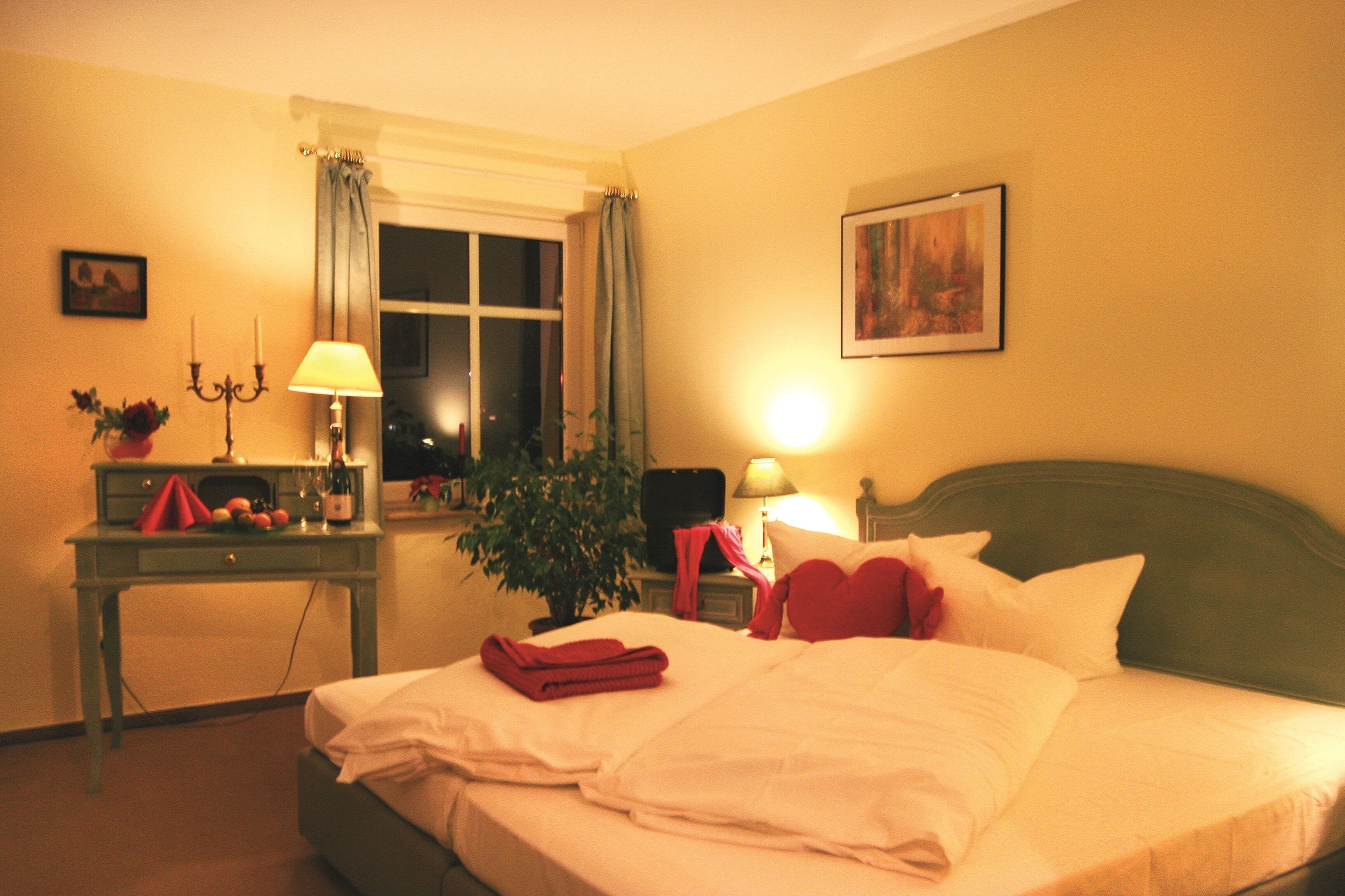 Una habitación de hotel con cama, escritorio con lámpara, planta y ventana con cortinas.
