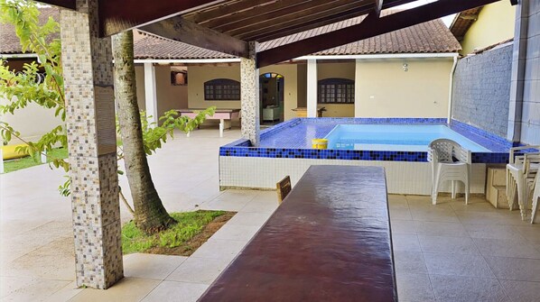 A covered outdoor area with a swimming pool, a long bench, and a small table.