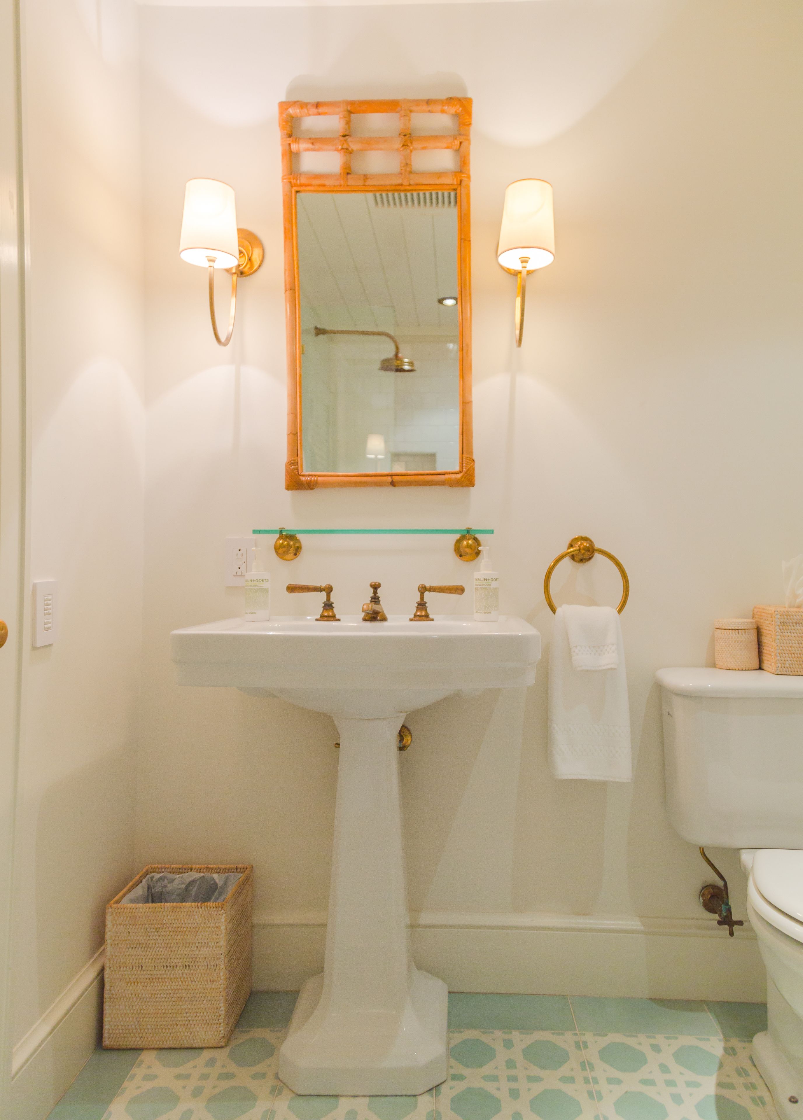 A bathroom with a pedestal sink, a mirror with a bamboo frame, two wall-mounted lights, a toilet, and a small wicker basket.