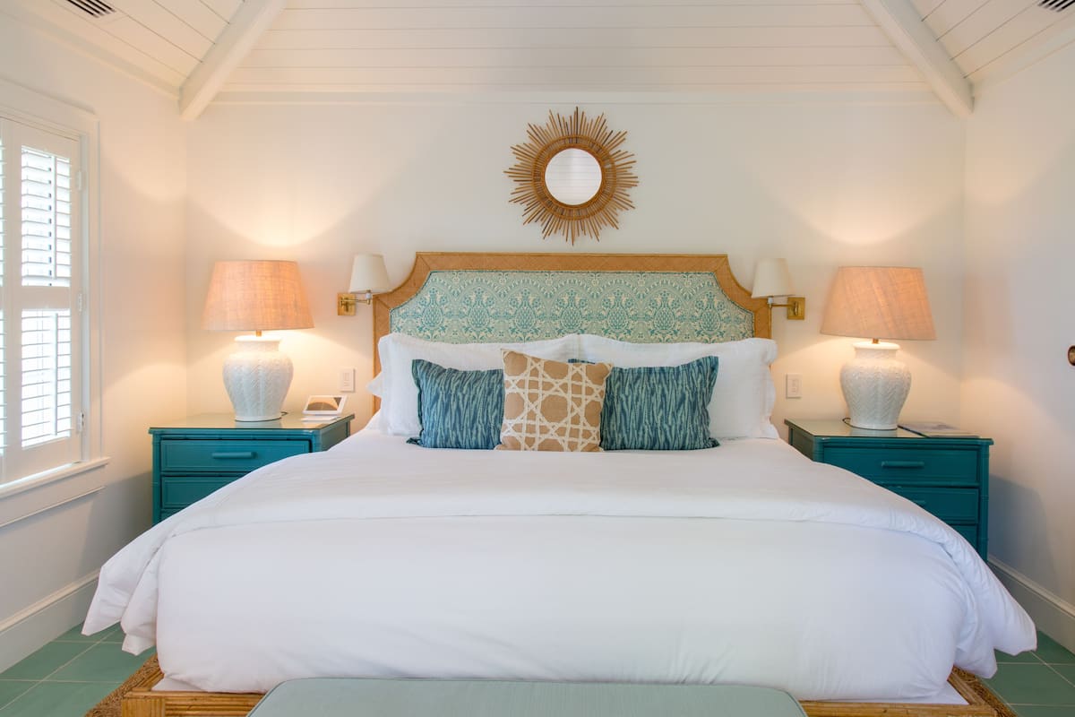 Una cama bien hecha con un cabecero decorativo, dos mesitas de noche con lámparas y una ventana con persianas.