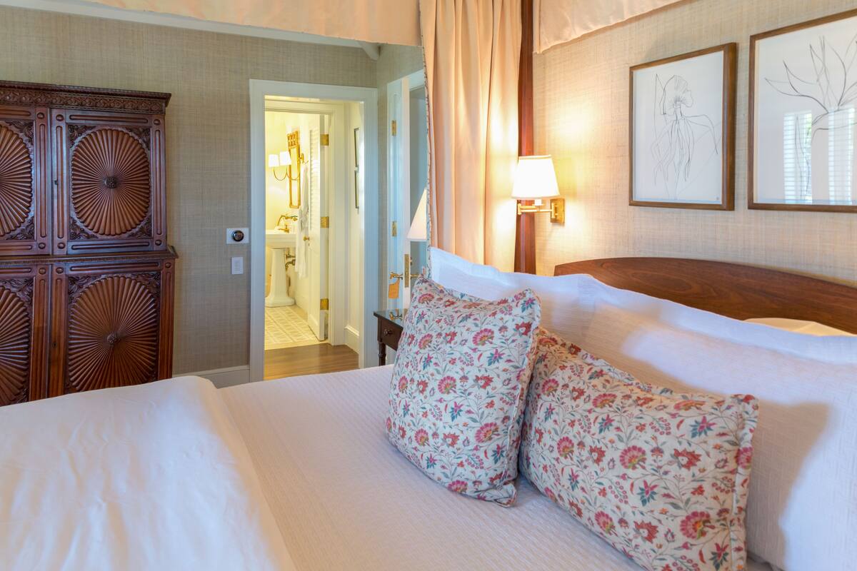 Una habitación de hotel con una cama grande, almohadas estampadas con motivos florales, un armario de madera y dos cuadros con ilustraciones botánicas en la pared.