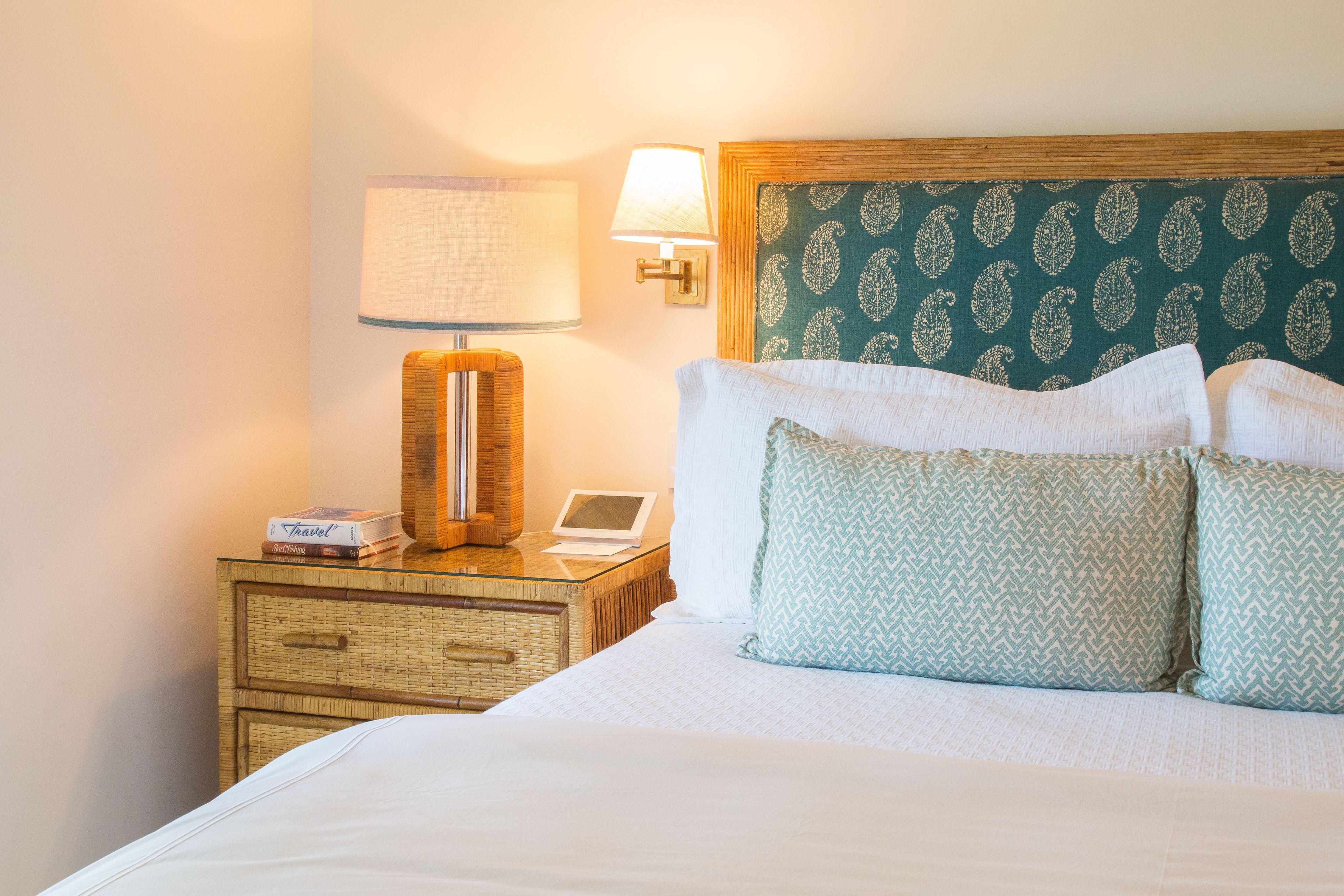 A neatly made bed with white linens and two patterned pillows, a bedside lamp, a small bedside table with a lamp, and a wall-mounted light fixture.