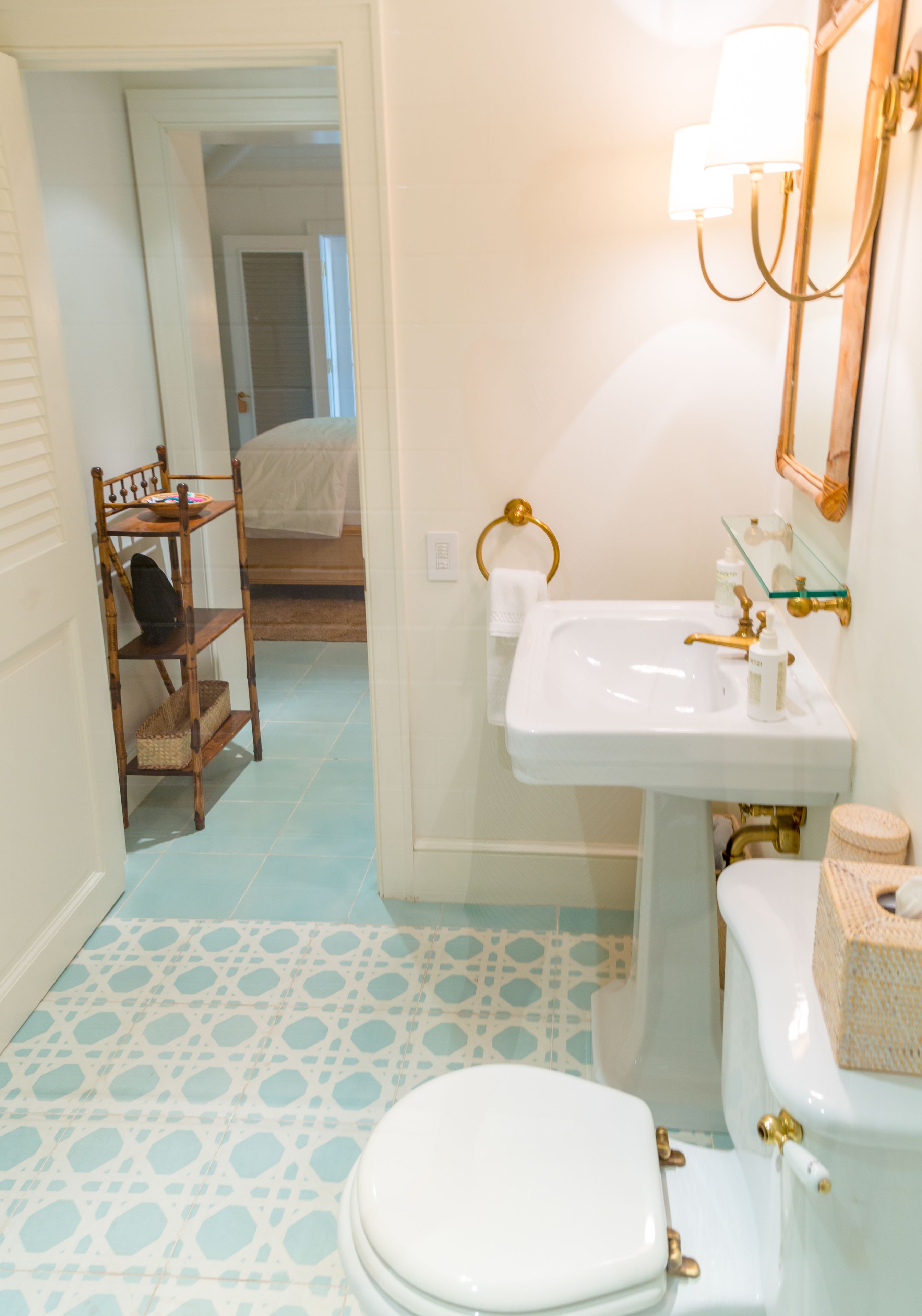 A bathroom with a white sink, toilet, and a mirror with a light fixture. There is a wooden shelf with baskets and a towel rack. The floor has a patterned tile design.