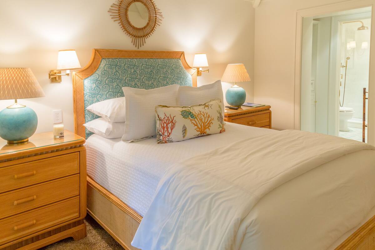 Una cama bien hecha con sábanas blancas y almohadones decorativos, flanqueada por mesitas de noche de madera con lámparas, en una habitación luminosa con vistas al baño.