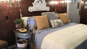 A bedroom with a wooden headboard, a round side table with a globe and a book, a bed with multiple pillows, and two wall-mounted chandeliers.