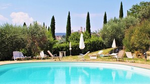 A swimming pool with lounge chairs and umbrellas, surrounded by greenery and tall trees, with a cityscape in the background.