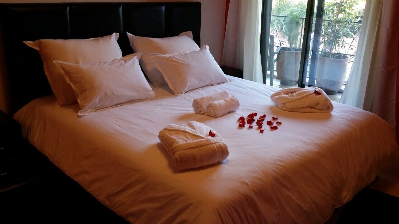 A neatly made bed with white linens and towels, a black headboard, and a view of potted plants through a window.