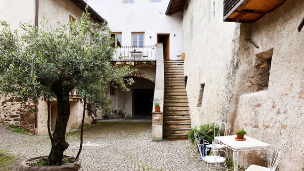 Ein Innenhof mit Kopfsteinpflaster, ein Blumentopf mit Baum, ein weißer Tisch mit Stühlen und eine Treppe, die zu einer oberen Ebene führt.