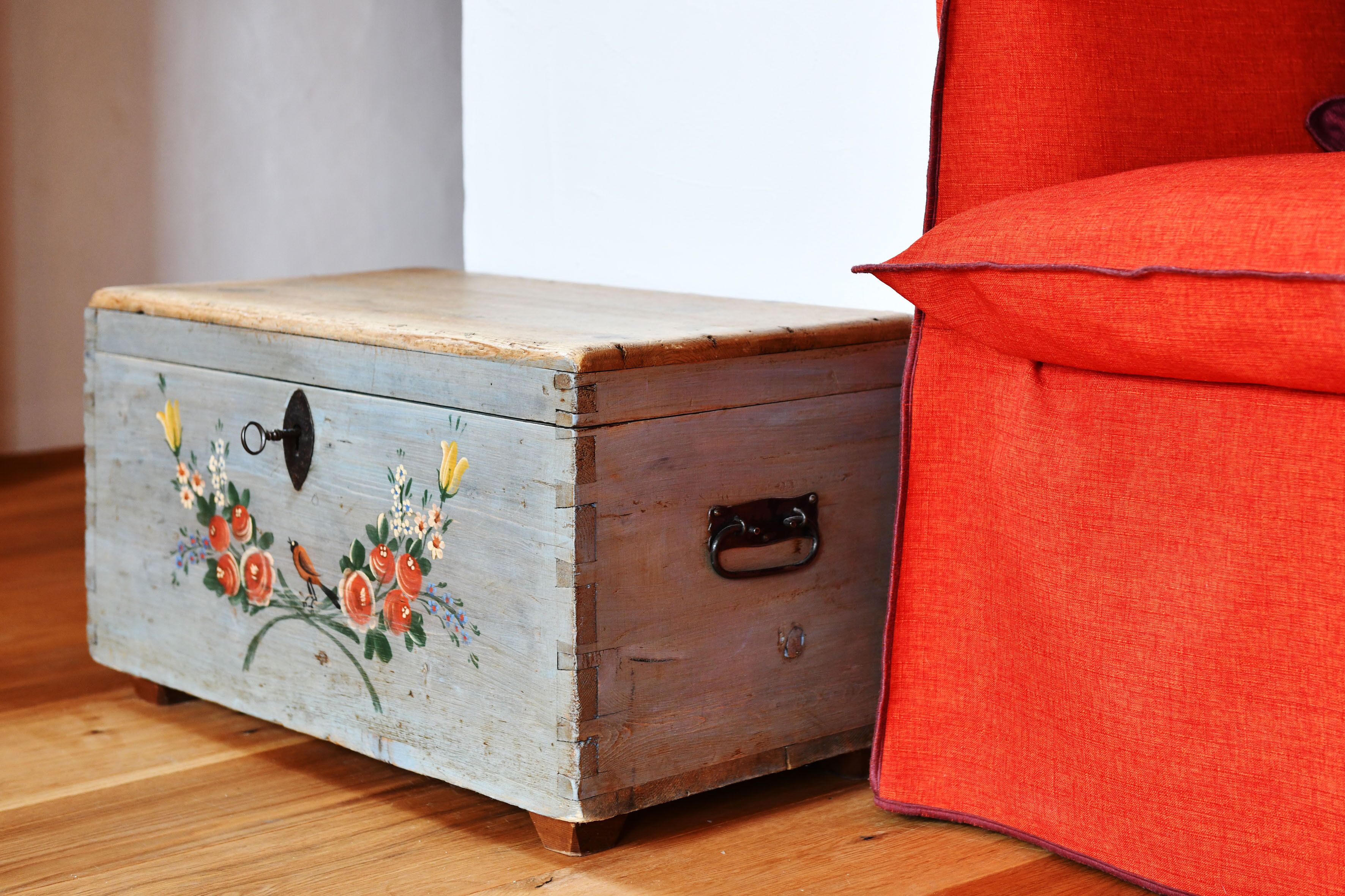 Une boîte en bois ornée de motifs floraux peints et dotée d’une anse en métal, posée sur un plancher en bois, à côté d’un fauteuil rouge recouvert de tissu.