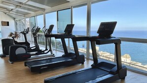 Un gimnasio con cintas de correr y equipo de ejercicio, ventanas amplias con vista al mar.