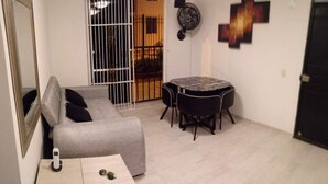 Un petit salon bien éclairé, avec un canapé gris, une petite table sur laquelle se trouve un téléphone, une table à manger ronde entourée de chaises et une porte coulissante à barreaux.