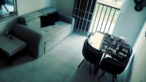 Un petit salon moderne avec un canapé gris, une petite table et une chaise. Une grande fenêtre avec des barreaux en métal offre une vue sur l’extérieur.
