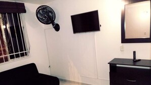 Une pièce avec un canapé noir, un téléviseur fixé au mur, un petit meuble et une fenêtre avec des barreaux.