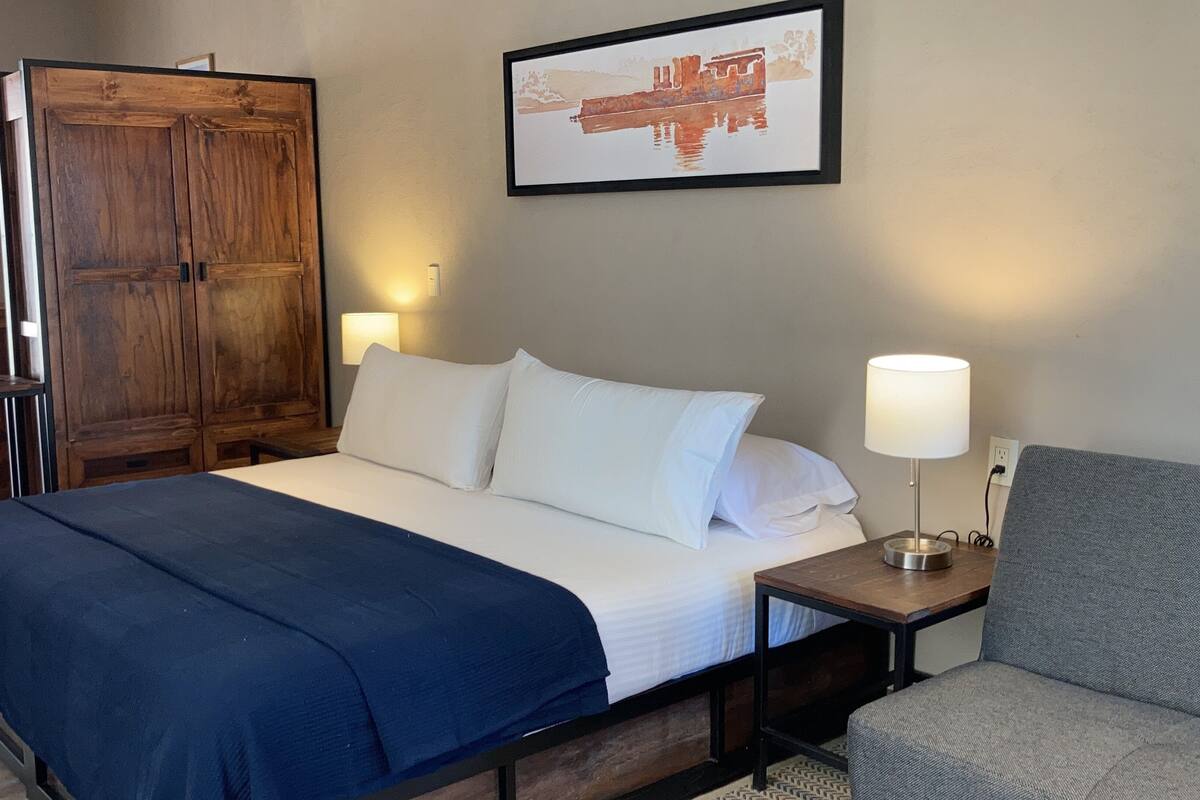 Habitación de hotel con una cama grande, un armario de madera, un cuadro enmarcado en la pared y un escritorio con lámpara.