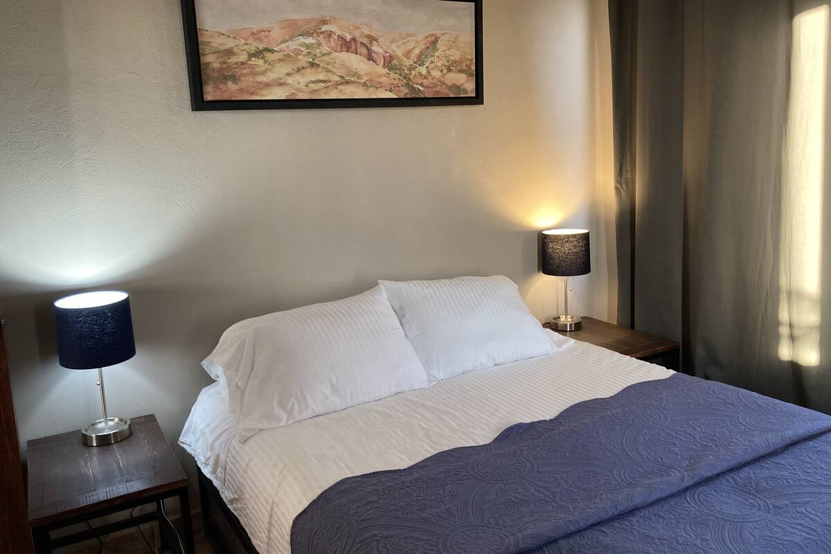 Una cama cuidadosamente tendida con sábanas blancas y una colcha azul estampada, flanqueada por dos mesitas de noche con lámparas, una a cada lado. Un cuadro con paisaje enmarco cuelga sobre la cama.