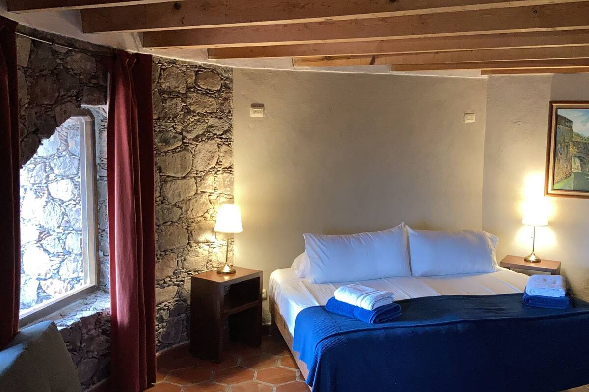 Un dormitorio con pared de piedra, una cama con sábanas blancas, dos lámparas de noche, una silla y un cuadro en la pared.