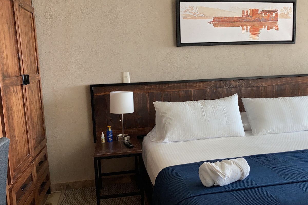 Una cama bien hecha con almohadas blancas y una colcha azul, un cabecero de madera, dos lámparas de noche, una mesita con una botella y un control remoto, y un cuadro en la pared.