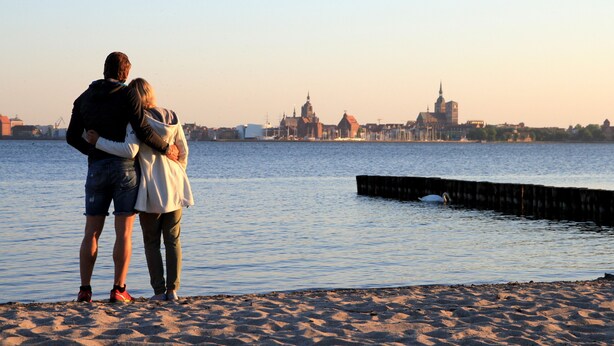 A couple embracing on a sandy beach with a cityscape in the background.