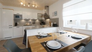 A modern kitchen with white cabinets, a wooden dining table set with plates, glasses, and cutlery, and a view of the outdoors through a window.