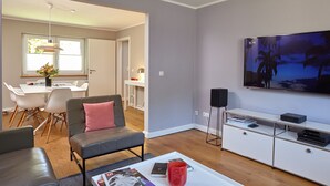 A modern living room with a grey sofa, a coffee table with magazines, a TV on the wall, and a dining area in the background. - 5 * Holiday home Düneneck 10 - Holiday home Düneneck 10 (Sankt Peter-Ording)