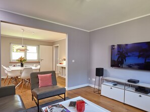 A modern living room with a grey sofa, a coffee table with magazines, a TV on the wall, and a dining area in the background.