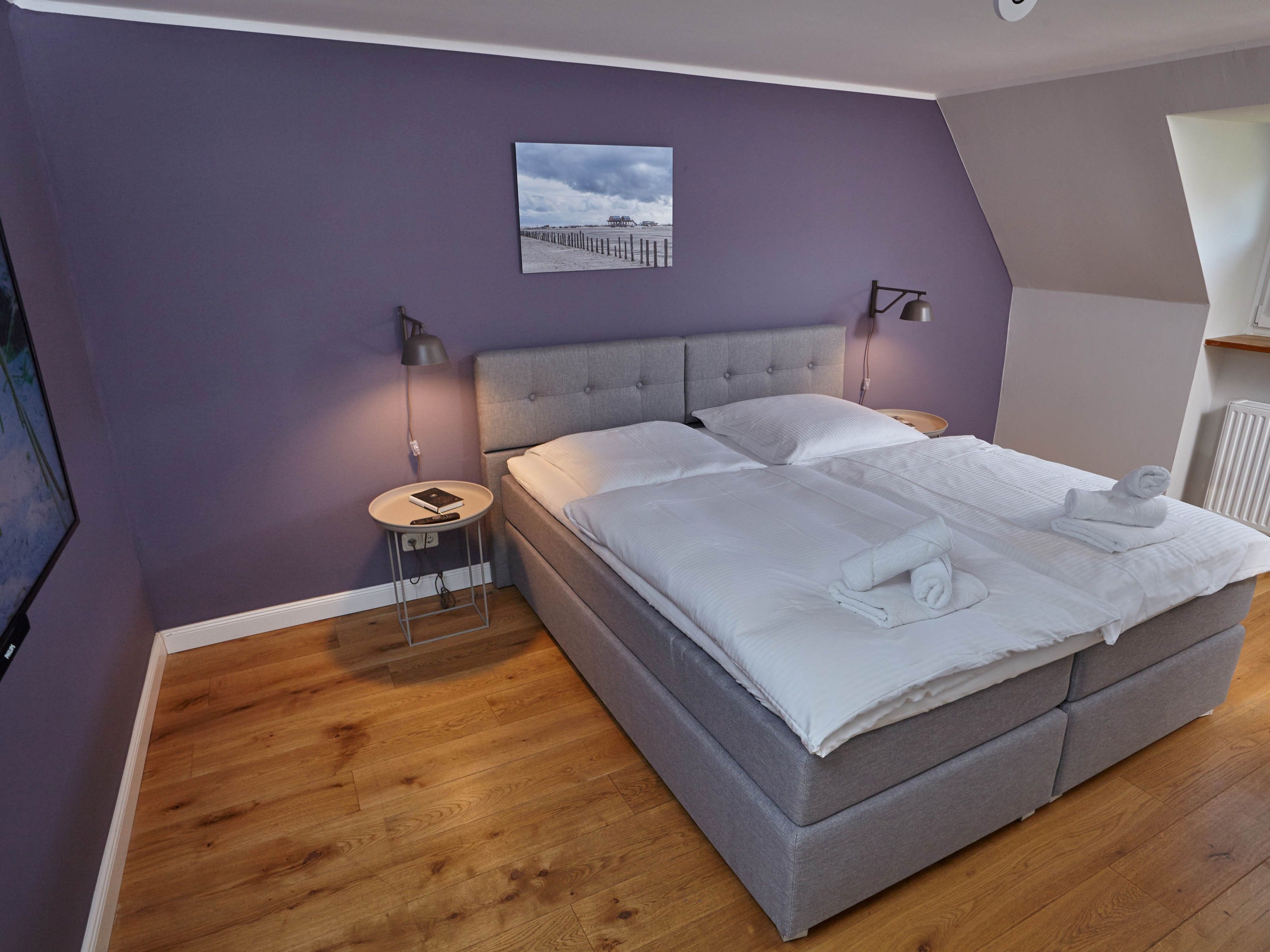 A bedroom with a grey bed, wooden flooring, a nightstand with a lamp, and a framed picture on the wall.
