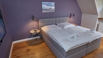 A bedroom with a grey bed, wooden flooring, a nightstand with a lamp, and a framed picture on the wall.