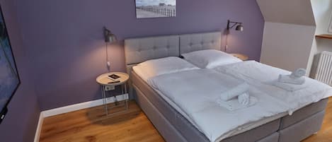 A bedroom with a grey bed, wooden flooring, a nightstand with a lamp, and a framed picture on the wall.