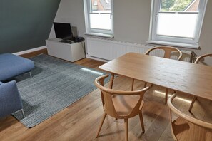 A room with a dining table, four chairs, a blue sofa, a TV on a white cabinet, and a radiator.