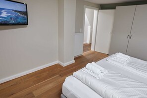 Une chambre avec un grand lit, des murs blancs, un plancher en bois, un téléviseur fixé au mur et une vue sur la mer par la fenêtre.