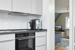 A modern kitchen with white cabinets, a built-in oven, a coffee maker, and a television in the adjacent room. - Apartment for 4 persons Düneneck 6 - Apartment for 4 persons Düneck 6 (Sankt Peter-Ording)