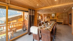 Un coin repas en bois avec une longue table dressée pour le repas, un plaid en fourrure sur une chaise et une grande fenêtre donnant sur l’extérieur.