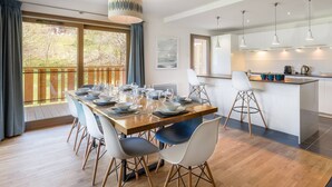 A modern dining area with a wooden table set for six, surrounded by white chairs with black accents. A kitchen with white cabinets and a bar area is visible in the background. - Luxury flat for 8 people - modern, close to the centre and slopes (CHATEL)