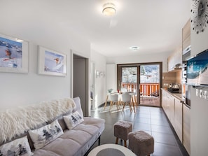 Ein modernes Wohnzimmer mit einer Couch, einem Essbereich und einer Küche. An der Wand befinden sich gerahmte Bilder, und es gibt einen Balkon mit Blick auf die verschneite Landschaft.