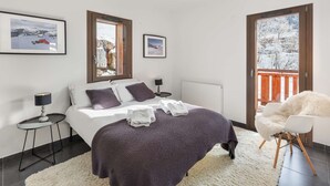 A modern bedroom with a bed, two bedside tables, a chair, and a view of a snowy landscape through the windows. - Modern, cosy 4-person flat - close to the centre and skiing (Châtel)