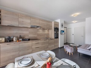 A modern kitchen with wooden cabinetry, a dining area with a table set for two, and a living area with a sofa and ottoman.
