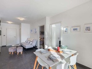 Ein modernes Wohnzimmer mit einer Couch, einem Esstisch für zwei Personen und einem durch eine offene Tür einsehbaren Küchenbereich.