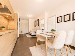 A modern kitchen with wooden cabinets, a dining area with a white table and fluffy chairs, and a living room with a sofa and a small side table.
