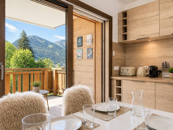A modern kitchen with wooden cabinets, a dining area with a table set for two, and a view of mountains through an open door.