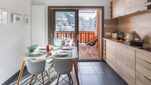 A modern kitchen with wooden cabinets, a dining area with a table set for two, and a view of a snowy deck outside. - Modern, cosy 4-person flat - close to the centre and skiing (Châtel)