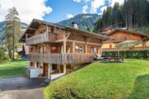 A wooden chalet with a balcony, a covered outdoor seating area, and a view of the surrounding mountains.